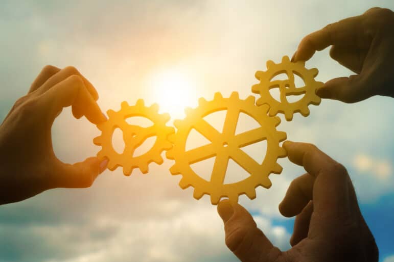 Three hands of businessmen assemble gears into a puzzle in a single whole against the backdrop of the sunset
